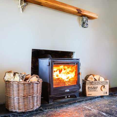 Wood-burning stove with fire lit, surrounded by a basket of logs and a box of Ecofuel birch wood bundles.