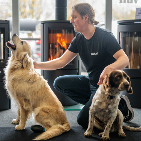 Janis Vitols, founder of ECOFUEL™, with two dogs beside a wood stove — symbolising warmth, connection, and sustainable living.