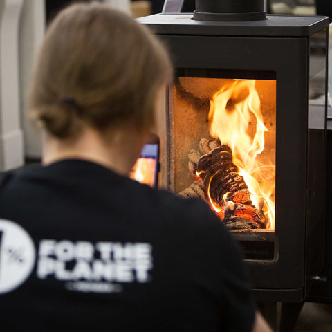Janis the Ecofuel founder wearing a black shirt with '1% for the Planet' logo, looking at a wood stove with flames.