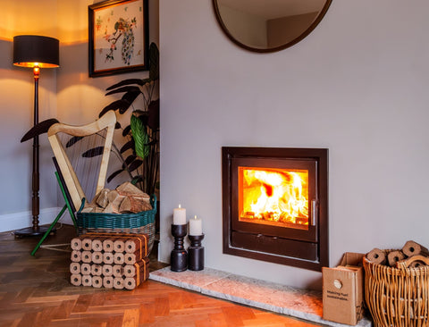 A modern wood-burning fireplace with a dark metal frame and glowing fire, set in a styled room with a stone hearth, candles, stacked ECOFUEL firewood, a floor lamp, and a wicker basket.